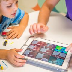 Two children sitting at a table are playing and interacting with a tablet.